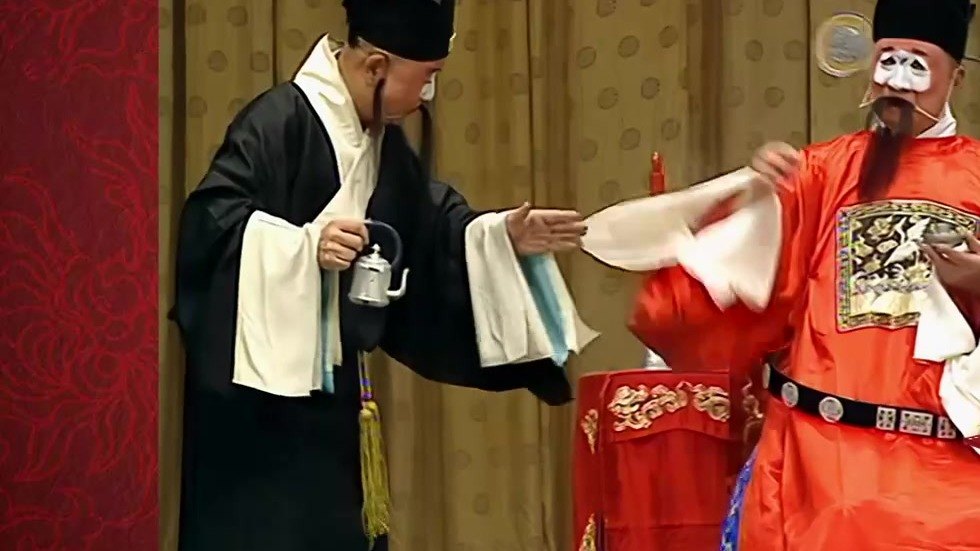 京剧《审头刺汤》「审头」公堂录像截帧：老生（左）与汤勤（丑，右，白面红官衣）当堂对白