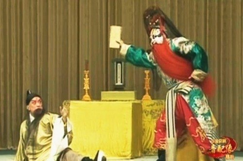 京剧《打金砖》剧照：太庙一场，供桌设神主牌位与烛台，一角色跌坐惊退，净角（红黑脸谱、红满髯、绿蟒）立而举牌（央视网《京剧音配像〈打金砖〉》文内所载）