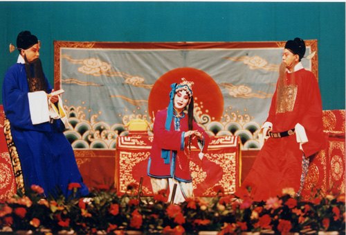 京剧《玉堂春》剧照：三堂会审一场，苏三（罪衣、鱼枷）与众官员同台（国家京剧院官网剧目页所载）