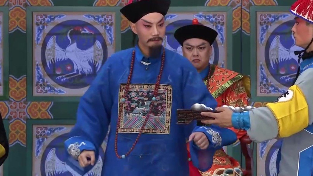 京剧《瘦马御史》演出照：差役托盘呈银锭、御史（蓝官衣、朝珠、黑三髯）伸手探看，红蟒官员在后——查库一节官场场面