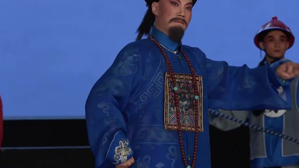 京剧《瘦马御史》演出照：老生扮御史（宝蓝官衣、补子、朝珠、黑三髯）作指认身段，身后差役牵铁链