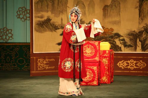 京剧《穆桂英挂帅》剧照：穆桂英着团花红蟒、持手帕立于桌案前（国家京剧院官网剧目页所载）