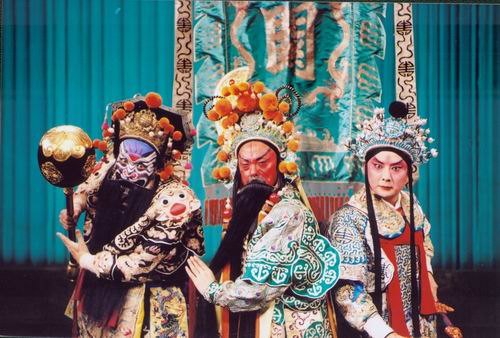 京剧《群英会》剧照：三人同场亮相——左为勾脸净角（黑髯、绒球珠饰硬盔、黑底金绣戏衣，持金色瓜形砌末），中为红脸长髯角色（绿底金绣戏衣），右为俊扮无髯年轻男角色（浅色绣花戏衣、系红带）；背景为湖蓝幕布与绘龙纹竖幅守旧（国家京剧院官网剧目页所载，未标注场次）