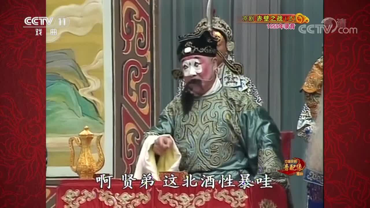 CCTV-11《中国京剧音配像精粹》京剧《赤壁之战》（上，1959年录音）播出画面：丑角扮相人物坐于桌案后举壶劝酒，旁立扎靠武将，底部字幕“啊贤弟 这杯酒性暴哇”