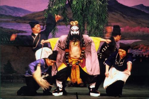 京剧《野猪林》剧照：鲁智深（净，勾脸、挂佛珠、着僧衣）居中，众角色环侍（国家京剧院官网剧目页所载）